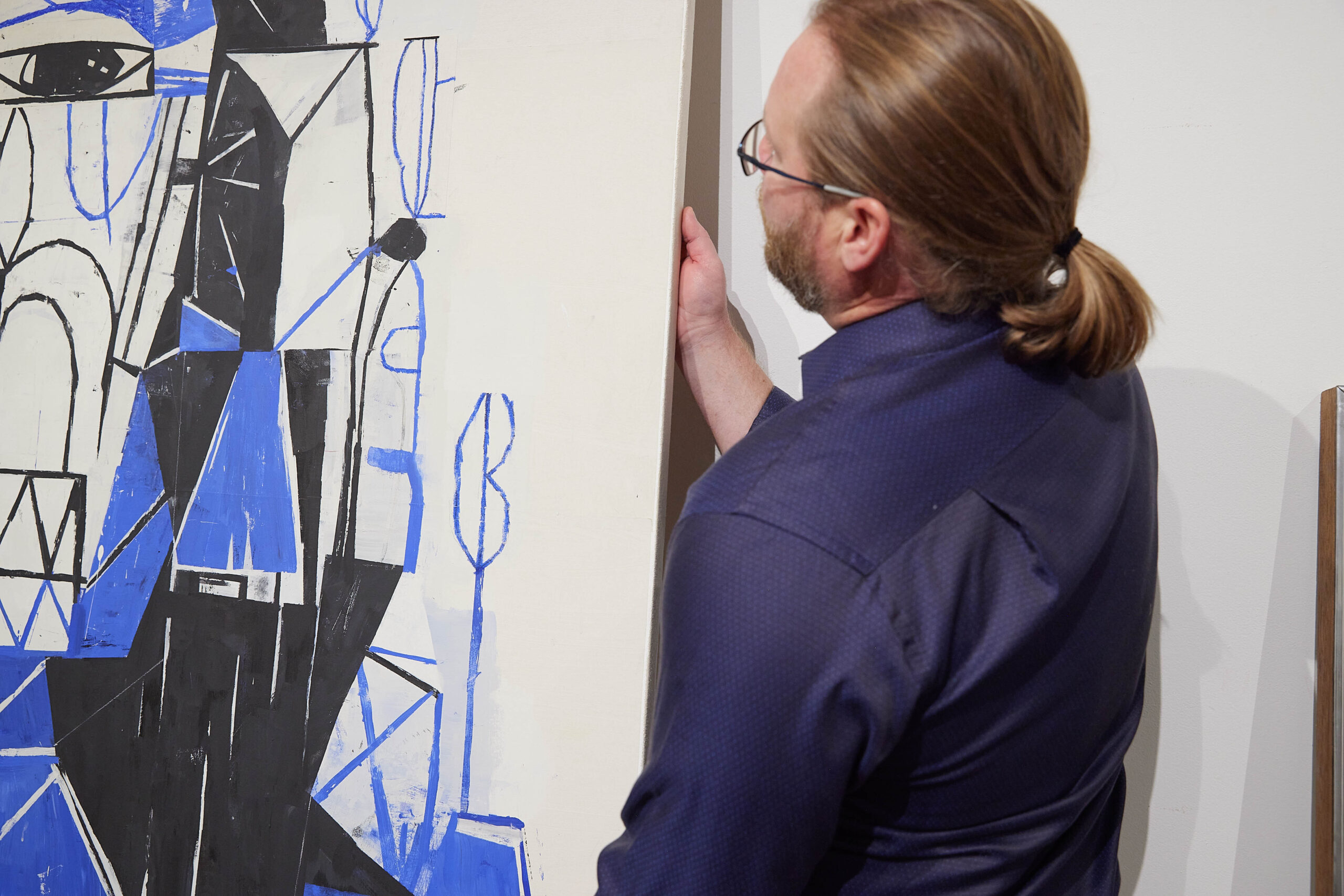 A man with a ponytail hangs an abstract artwork on a wall