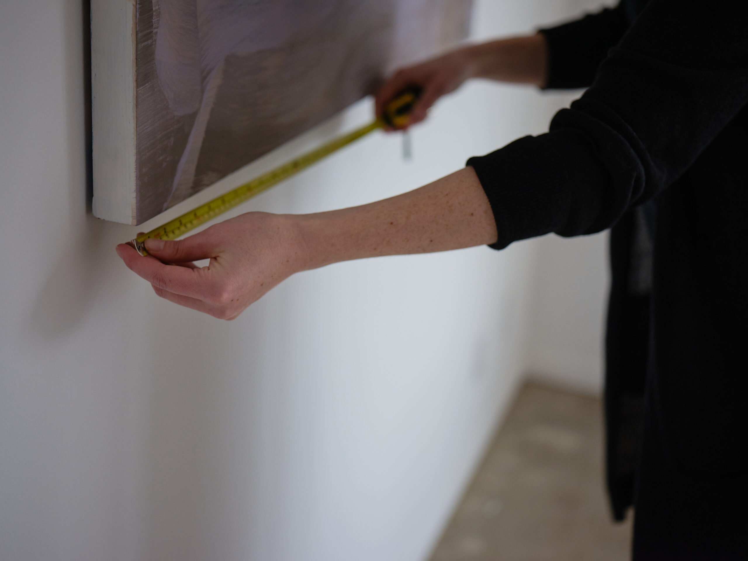 A woman uses a tape measure to measure an artwork hanging on the wall